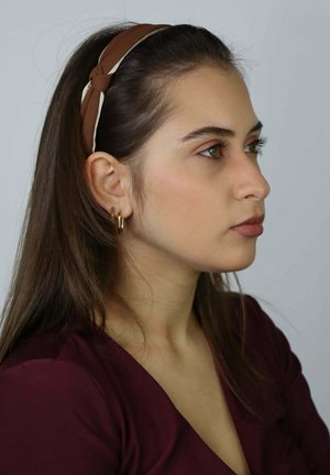 Brązowa opaska na głowę z tkaniny z węzłowym wzorem i kremowym akcentem. Modelka ma długie włosy, nosi złote kolczyki koła oraz bordową bluzkę.