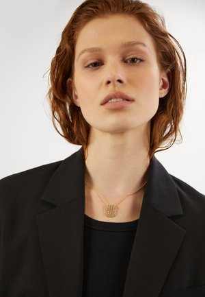 Gold-tone necklace with a rectangular pendant featuring multiple sparkling stones, worn over a black top and under a black blazer.
