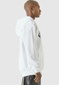 Hoodie blanc en coton, avec une poche avant, des poignets côtelés, et un design graphique noir sur la poitrine et la manche.