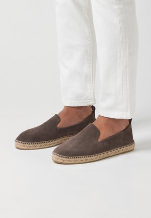Hnědé semišové espadrilky s zakřiveným designem na obutí, juteovou podrážkou a malým podpatkem, kombinované s bílými džíny.