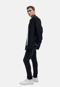 Camisa de mezclilla negra sobre una camiseta blanca, combinada con pantalones ajustados negros y zapatillas blancas. La camisa tiene un corte relajado y un bolsillo en la parte delantera.