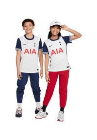 Zwei Kinder posieren in Sportbekleidung: ein weißes Hemd mit marineblauen Akzenten und einem roten "AIA"-Logo, kombiniert mit dunkelblauen und roten Jogginghosen.
