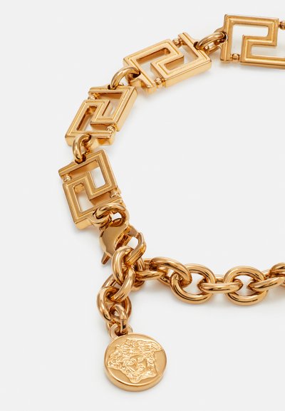 Bracelet en chaîne dorée présentant des maillons géométriques avec un motif en relief et un charme rond illustrant un design complexe. Fini lisse et brillant.