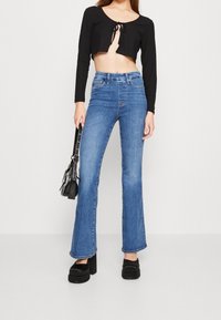 Svart ribbad croptop med knopplås, matchad med ljusblå utsvängda jeans och svarta plattformsskor. Liten svart handväska i handen.