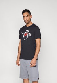 Μαύρο βαμβακερό T-shirt με πολύχρωμο γραφικό δεινοσαύρου που φοράει κόκκινη φανέλα, σετ με γκρι σορτς που εμφανίζουν μαύρο λογότυπο.