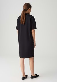 Schwarzes Kleid mit kurzen Ärmeln und einem Kragen, mit einem geraden Schnitt und einer glatten Textur. Getragen mit schwarzen flachen Schuhen.