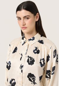 Cream blouse with black floral print, featuring a button-down front, collared neck, and softly gathered sleeves for added texture.