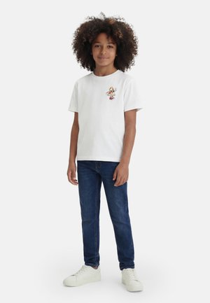 Kind mit lockigem Haar, das ein weißes T-Shirt mit einem Cartoon-Pizzastück, blaue Jeans und weiße Sneakers trägt, steht vor einfachem Hintergrund.
