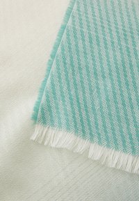 Textile scarf featuring a herringbone pattern in teal and white, with frayed edges, layered over a smooth, cream-colored fabric.