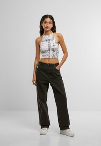 Fehér pánt nélküli crop top, fekete paisley mintával; laza, sötét farmernadrággal párosítva. A modell egy világosszürke háttér előtt áll.
