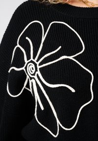 Zwarte gebreide trui met een groot wit geborduurd bloemenpatroon op de voorkant. De stof heeft een geribbelde textuur.