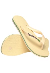 Des tongs de couleur crème avec des semelles texturées et un semelle intérieure à motifs. Les brides entre les orteils présentent un logo et un détail du drapeau brésilien.