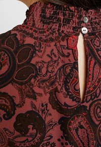 Blusa marrón con estampado y cuello alto fruncido, que presenta una abertura en la parte posterior con forma de ojal y dos botones. Los patrones incluyen diseños de paisley.