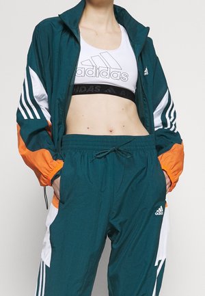 Person wearing teal Adidas track jacket and pants with orange and white accents, white Adidas sports bra, hands in pockets.