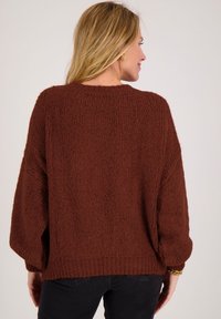 Vrouw met blond haar, goudkleurige oorringen en een grof gebreide bruine trui, gezien van achteren tegen een effen achtergrond.