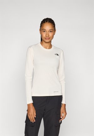 Donna con capelli intrecciati che indossa una maglia bianca a maniche lunghe North Face e pantaloni cargo neri, in piedi su uno sfondo chiaro e uniforme.