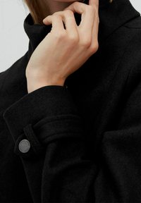 Cappotto di lana nera con colletto alto, caratterizzato da un polsino con bottone e tessuto testurizzato. Primo piano di una mano che regola il colletto.