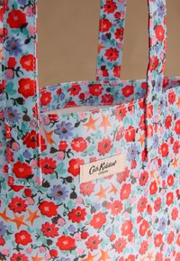 Mėlynos šviesios spalvos raštuotas audinio pirkinių maišelis su raudonais žiedais, žaliais lapais ir oranžinėmis žvaigždėmis. Ant maišelio siuvinėta etiketė su užrašu „Cath Kidston London.“