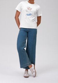 Bílá tričko s grafickým designem znázorňující postavu a květiny, kombinované s širokými modrými džíny a šedobílými teniskami.