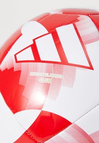 Raudona ir balta futbolo kamuolys su geometriniu raštu, ant kurio auksinėmis raidėmis parašyta "MATCH BALL REPLICA CLUB". Lygus, blizgus paviršius.