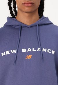 Blå sweatshirt med huva, prydd med vit text "NEW BALANCE" och en orange logotyp. Mjuk väv med avslappnad passform och justerbara snören.