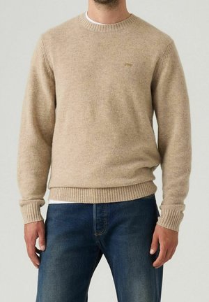 Homme portant un pull en laine beige col rond et un jean bleu, debout devant un fond clair uni.