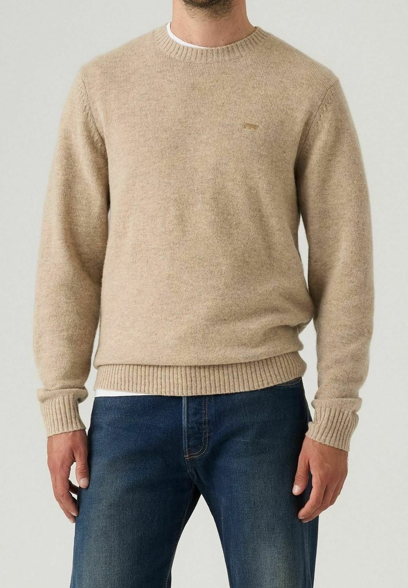 Homme portant un pull en laine beige col rond et un jean bleu, debout devant un fond clair uni.