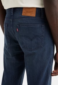 Zadní pohled na osobu v tmavě modrých džínách Levi's s koženou nášivkou a červenou značkou Levi's na pravé zadní kapse.