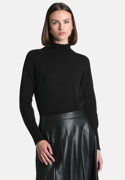 Femme aux cheveux bruns mi-longs portant un pull noir à col roulé et une jupe plissée en cuir noire, debout devant un fond uni.