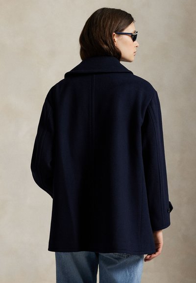 Naine kannab tumel sinist mantel ja päikeseprille, seisab külje pealt, helepruuni juukse ja helesiniste teksadega neutraalsel taustal.