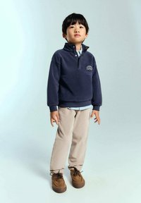 Pulover din fleece de marină cu fermoar până la jumătate și logo; cămașă cu guler de culoare albastru deschis dedesubt; pantaloni bej; pantofi maro. Copilul stă în fața unui fundal deschis.