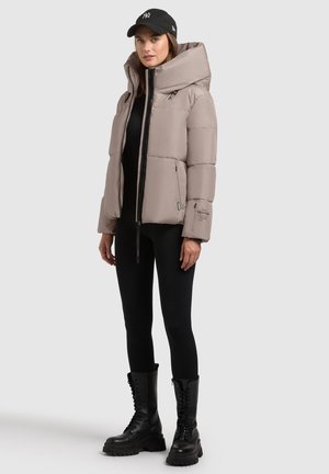Puffjacke in hellem Taupe mit hohem Kragen, Reißverschluss vorne und zwei seitlichen Taschen. Getragen über einem schwarzen Oberteil und kombiniert mit schwarzen Schnürstiefeln.