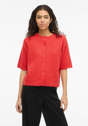 Femme aux cheveux bouclés portant un cardigan rouge à manches courtes avec des boutons et un pantalon noir, debout devant un fond blanc.