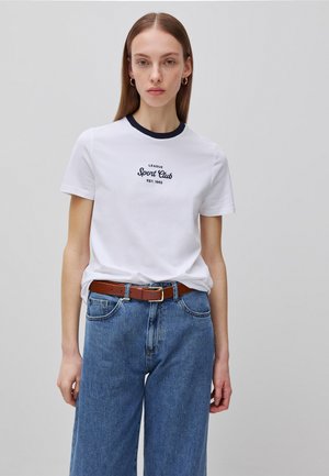 Junge Frau mit glattem braunen Haar trägt ein weißes "Sport Club" T-Shirt, blaue Jeans und braunen Gürtel, steht vor einem schlichten grauen Hintergrund.