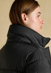 Černá péřová bunda s vysokým límcem, hladkou texturou látky, viditelným švem a zipovým zapínáním. Má štíhlou siluetu.