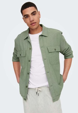 Jeune homme portant une chemise vert clair boutonnée sur un T-shirt blanc avec un pantalon beige rayé, regardant vers la caméra les mains derrière le dos.