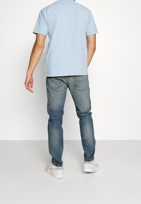 Světle modré bavlněné tričko, krátké rukávy, detail loga na zádech, kombinováno s slim-fit vybledlými modrými džíny a bílými teniskami. Jednoduché pozadí.