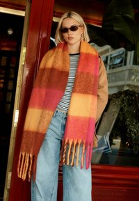 Wollschal mit auffälligem Streifenmuster in Orange, Pink, Braun und Gelb, mit fransigen Enden. Das Modell trägt ein gestreiftes Oberteil und eine Jeans.
