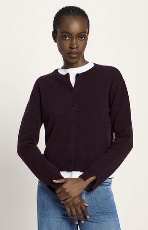 Eine Frau mit geflochtenem Haar, die einen dunkelvioletten Knopf-Cardigan über einem weißen Shirt und blauen Jeans trägt, steht mit verschränkten Armen.