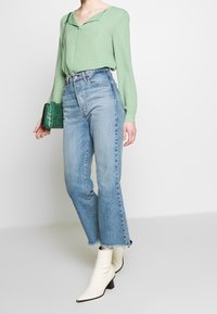 Blusa verde a maniche lunghe con scollo a v, abbinata a jeans azzurri a vita alta con orlo corto e sfrangiato. Stivaletti bianchi alla caviglia e borsa verde.