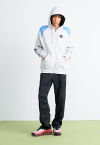Sweatshirt com fecho éclair cinza claro com destaques azuis, apresentando um logo no peito, combinado com calças de atletismo pretas e sapatilhas desportivas com padrão.