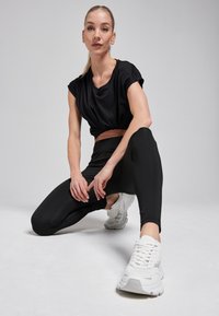 Zwart cropped sporttop gecombineerd met zwarte leggings, met een gladde textuur. Witte sneakers met een gestructureerde zool, geplaatst op een lichte achtergrond.