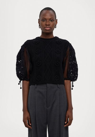 Černý pletený crop top s texturovaným květinovým vzorem a průsvitnými balónovými rukávy. Vytvořeno s šedými kalhotami s vysokým pasem, zdůrazňujícími moderní siluetu.