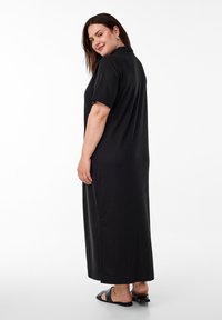 Abito lungo nero a coste con maniche corte e colletto, caratterizzato da una vestibilità rilassata. La modella sta in piedi, indossando sandali con tacco piatto.
