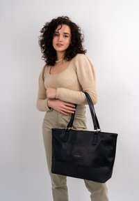 Black leather tote bag with two long handles, smooth texture, and gold branding detail on the front. Simple rectangular shape.