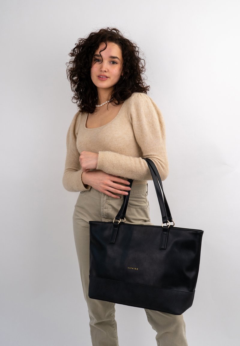 Black leather tote bag with two long handles, smooth texture, and gold branding detail on the front. Simple rectangular shape.