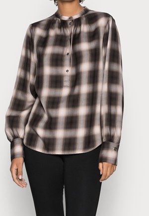 Persona che indossa una camicetta ampia a maniche lunghe a quadri neri e beige, con bottoni al colletto e ai polsini, abbinata a pantaloni neri.