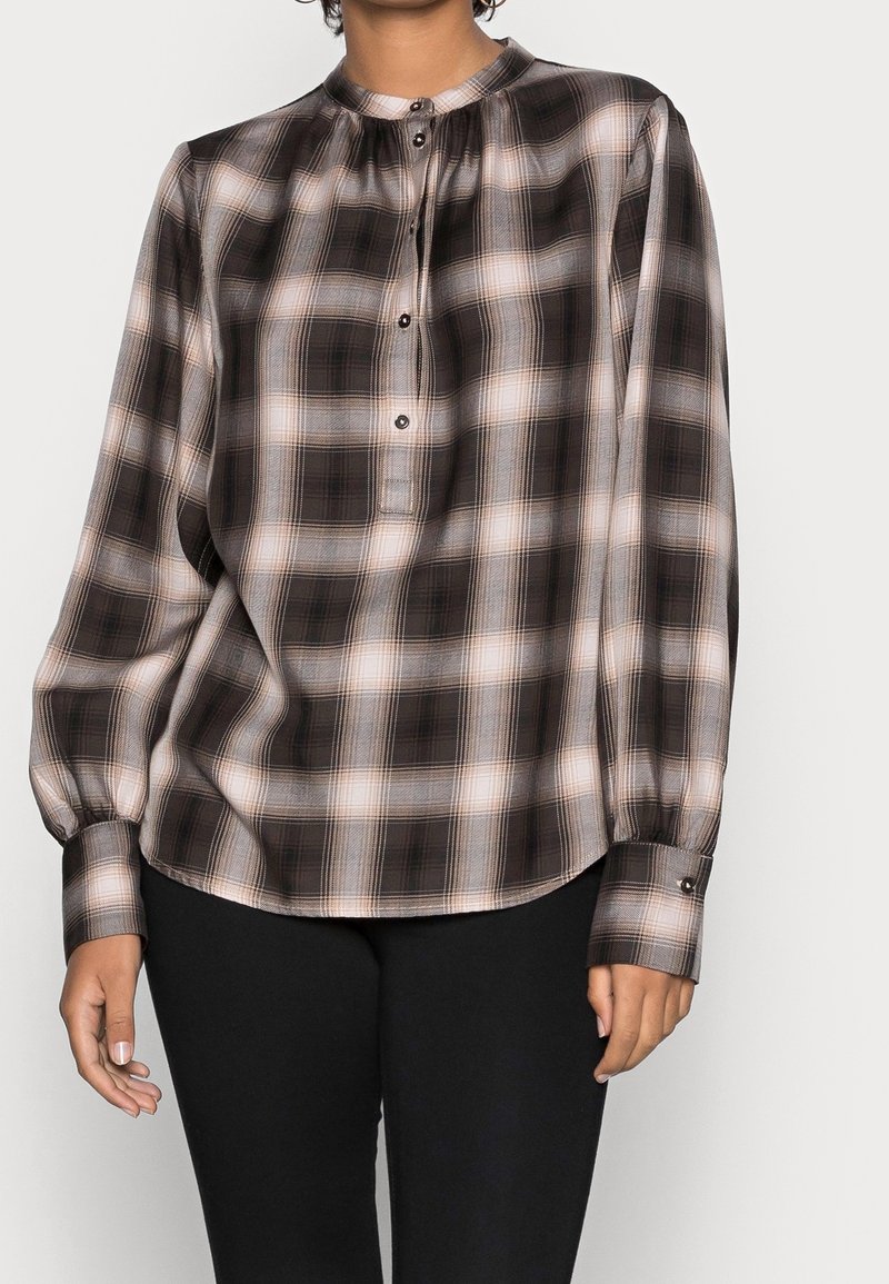 Personne portant une blouse ample à manches longues, à carreaux noirs et beiges, avec des boutons au col et aux poignets, associée à un pantalon noir.