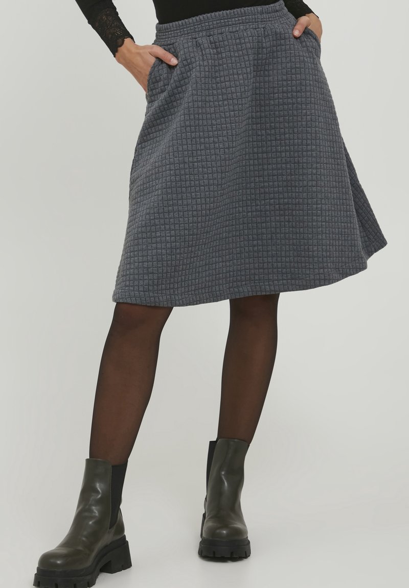 Jupe grise texturée, longueur genoux, avec un motif à carreaux, dotée d'une taille élastique et de poches latérales. Associée à des bottines noires.