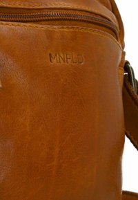 Bruine leren tas met een gladde textuur en een ritsvak. Heeft een gegraveerd "MNFLD" logo op het oppervlak. Verstelbare schouderriem zichtbaar.